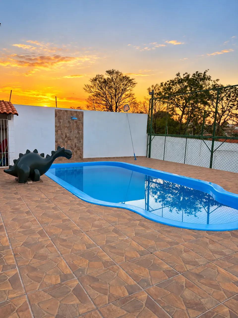 Piscina azul com deck amplo e um dinossauro decorativo, com o céu alaranjado do pôr do sol ao fundo