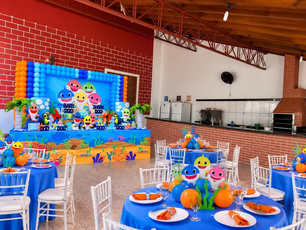 Salão coberto com parede de tijolinho, painel de balões azul e laranja, mesas redondas azuis e cadeiras brancas, com a cozinha ao fundo